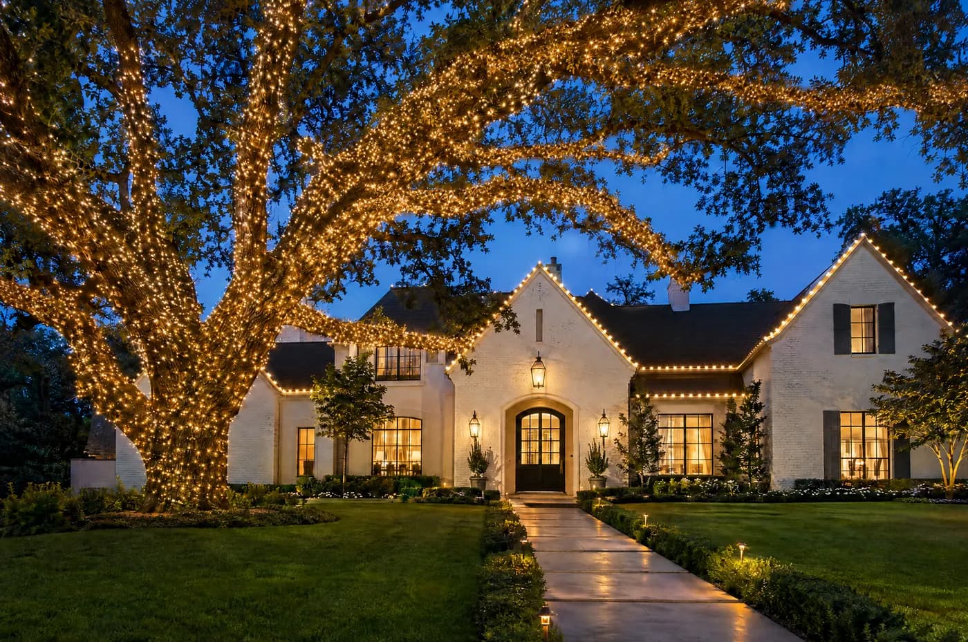 White brick and stucco home under a live oak canopy densely wrapped in warm white lights